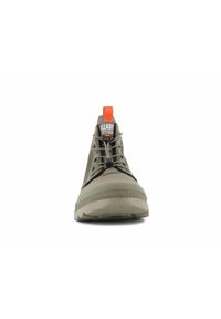 Vista frontal de una bota Palladium en verde oliva con un tirador naranja y cordones elásticos, sobre un fondo blanco.