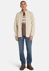 Beżowa bluza polarowa z wysokim kołnierzem, zamkiem błyskawicznym i dwoma bocznymi kieszeniami, nałożona na brązowy t-shirt z grafiką, zestawiona z niebieskimi dżinsami i brązowymi butami.