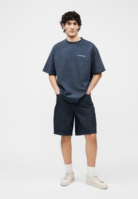 Jeune homme debout, les mains dans les poches, portant un t-shirt oversize bleu marine, un short noir à fines rayures, des chaussettes blanches et des baskets beige, sur fond blanc.