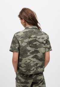 Camicia a maniche corte, corta, in motivo mimetico verde, realizzata in cotone. Presenta un colletto appuntito e un design a vestibilità ampia.
