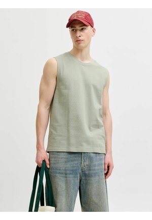 Noor mees rohelist varrukateta toppi, punast mütsi ja siniste teksapükstega, hoides valget käekotti tumeroheliste sangadega.