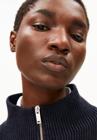 Nahaufnahme einer Person, die einen marineblauen gerippten Pullover mit hohem Kragen trägt, der mit einem silbernen Reißverschluss verziert ist, und darunter ein weißes Hemd.