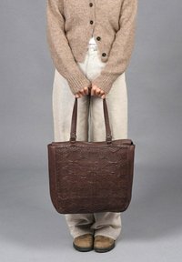 Persona con suéter beige, pantalones claros y zapatos marrones sostiene un gran bolso de cuero marrón oscuro y texturizado por las asas.