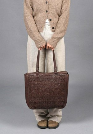 Persoon in een beige sweater, lichte broek en bruine schoenen houdt een grote, gestructureerde, donkerbruine leren tote-bag vast bij de handvatten.