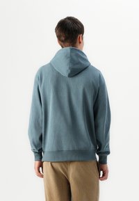 Sudadera de punto azul claro con una gran capucha, corte relajado y puños elásticos; combinada con pantalones de pana beige. Minimalismo en el diseño.