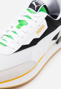 Baskets Puma blanches, noires et vertes avec logo jaune, lacets blancs, tissu en mesh, bout en suède et semelle en caoutchouc.