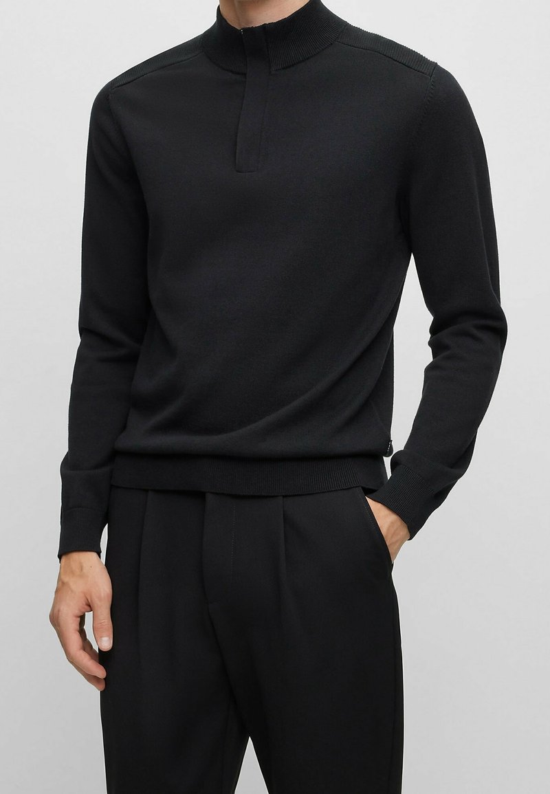 Man wearing a black long-sleeve knit pullover with a mock neck and concealed front placket, paired with black pleated trousers.