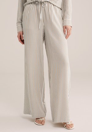 Kobieta ubrana w luźne, beżowe spodnie w pionowe białe paski z wysokim stanem i sznurkiem w pasie oraz białe sandały na odkryte palce.