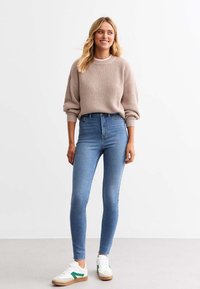 Suéter de punto marrón claro con mangas caídas, combinado con jeans ajustados azul de talle alto y zapatillas blancas con acentos verdes.