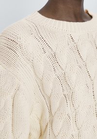 Pull en tricot couleur crème avec un motif en câble texturé, col rond et coupe ample avec des détails de couture visibles.