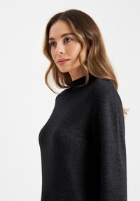 Zwarte high-neck trui van zacht, gestructureerd materiaal, met een relaxte pasvorm en lange mouwen, tentoongesteld tegen een effen witte achtergrond.