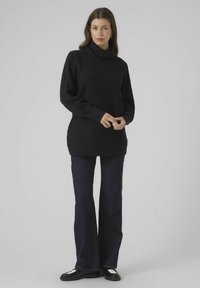 Maglione nero a collo alto lavorato a maglia con un motivo a chevron texturizzato, abbinato a jeans in denim scuro a campana e scarpe nere con dettagli bianchi.