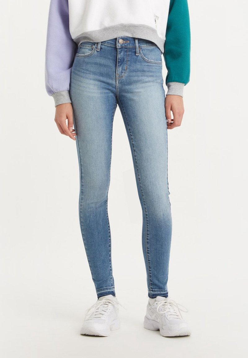 Personne portant un jean skinny bleu clair, un sweat-shirt color-block et des baskets blanches, debout devant un fond clair et uni.