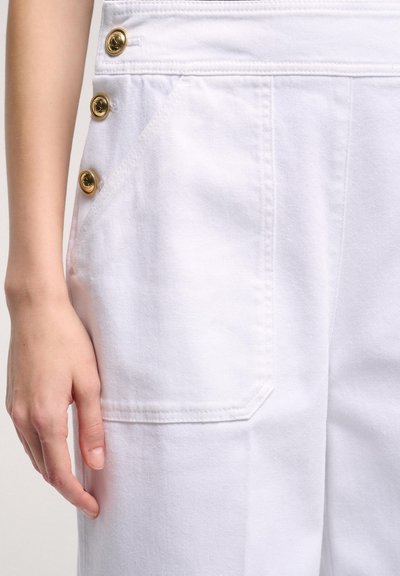 Salopette in denim bianco con tasca laterale, tre bottoni decorativi dorati e cuciture in evidenza. Tessuto liscio con una vestibilità strutturata.