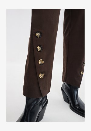 Bruine broek met een getailleerde pasvorm, voorzien van een rij decoratieve messing knopen langs de zoom en gecombineerd met zwarte hoge laarzen.