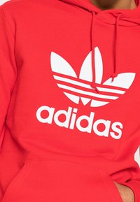 Röd hoodie i bomullsmaterial med en vit Adidas-logotyp och tre ränder. Inkluderar dragsnören och en framficka.