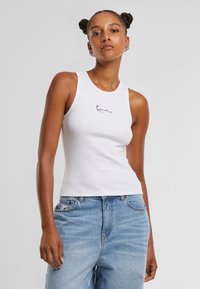 Débardeur blanc à côtes avec un col rond et un logo manuscrit noir sur la poitrine, associé à un jean en denim bleu clair.