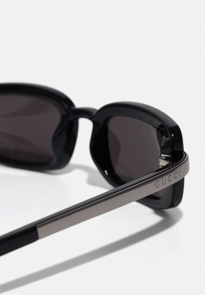 Lunettes de soleil noires avec une monture épaisse, des verres foncés et une branche grise élégante arborant l'inscription "GUCCI" gravée. Matériau en plastique lisse et brillant.