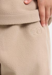 Pantaloni in felpa beige caratterizzati da una texture morbida, con un design ricamato di due faccine sorridenti sul lato vicino all'elastico in vita.