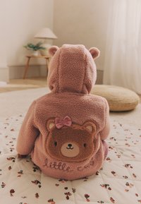 Różowa polarowa bluza z kapturem z uszami niedźwiedzia, z twarzą niedźwiedzia i kokardą na plecach, na tle wzorzystego koca i miękkich mebli.