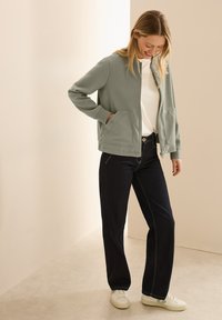 Sweatshirt zippé vert clair avec deux poches avant, associé à un jean en denim foncé et des baskets blanches. Tissu lisse, coupe décontractée.