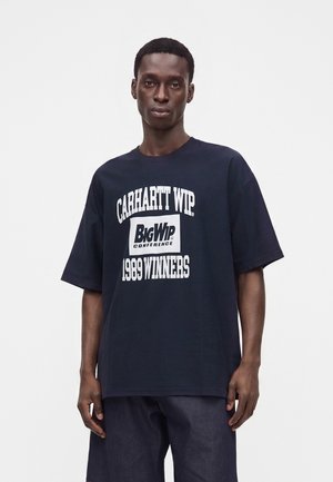 Mand der står og bærer en mørkeblå oversized T-shirt med hvid tekst, der lyder Carhartt WIP Big WIP Conference 1969 Winners.