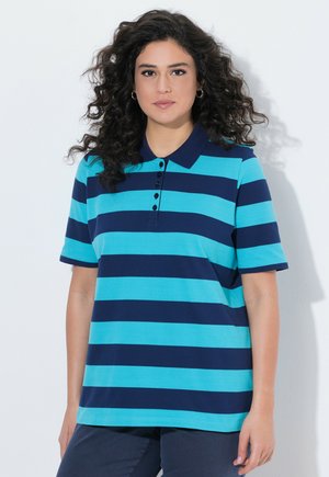 Kvinde med krøllet hår iført en blå og marineblå stribet kortærmet poloshirt, stående mod en ensfarvet hvid baggrund.