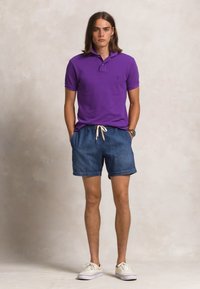 Lilla poloskjorte med krage, denimshorts med snøring og hvite joggesko. Modellen står med hendene i lommene mot en nøytral bakgrunn.