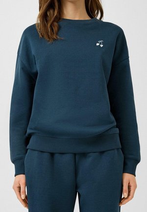 Femme portant un sweat-shirt bleu foncé et un pantalon assorti avec un petit motif blanc de cerise côté gauche de la poitrine, se tenant devant un fond uni.