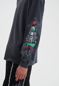 Langärmliges dunkelgraues Shirt mit bunten Grafikdesigns auf den Ärmeln, darunter ein Totenkopf und verschiedene florale Elemente.