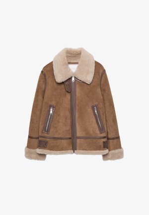 Femme portant une veste en daim marron avec doublure en fourrure crème et un jean bleu foncé, vue de dos sur un fond uni.