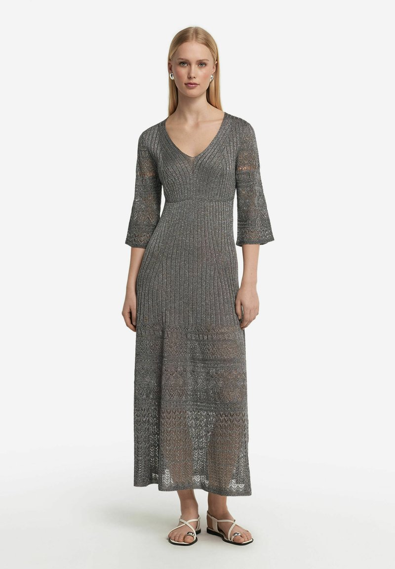Lång klänning med utflädd ärm i metallgrått trikåtyg, med V-ringning och texturerade mönster. Avslutas med en flödande längd som når anklarna.