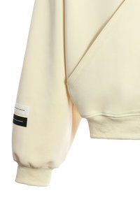 Sudadera color crema con puños y dobladillo acanalados, que presenta una costura diagonal única y una etiqueta de marca en la manga. Textura suave.