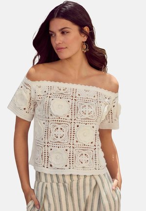 Haut en crochet blanc à épaules dénudées avec des motifs floraux, des bords festonnés et des manches courtes, associé à un pantalon en lin rayé.
