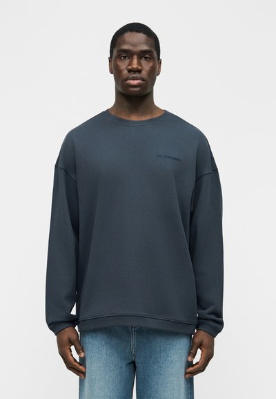 Άνδρας που φοράει σκούρο μπλε φούτερ με μακριά μανίκια και ανοιχτό μπλε τζιν, στέκεται μπροστά σε απλό λευκό φόντο.