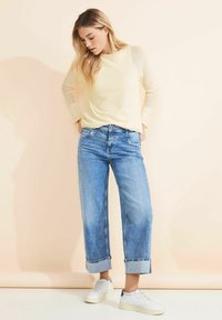 Helles gelbes langärmliges Oberteil mit Struktur, kombiniert mit hoch taillierten, hellblauen, weit geschnittenen Jeans, die an den Säumen aufgerollt sind, und weißen Sneakers mit dunkelblauen Akzenten.