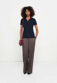 Marineblaues Polohemd mit Kragen, fünf Knöpfen und kurzen Ärmeln, kombiniert mit hoch taillierten, strukturierten braunen Hosen und einer lila Handtasche.