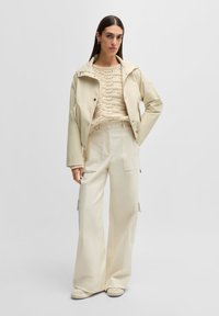 Jachetă scurtă din crem deschis, cu capse, asortată cu un pulover texturat din tricot crem și pantaloni evazați cu talie înaltă. Material neted, design minimalist.