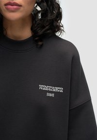Black sweatshirt with a high neckline and loose fit. Features a white logo “WRSTBHVR [030]” on the chest. Soft, textured fabric.