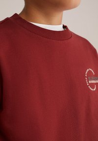 Burgundy sweatshirt med rund halsringning, med en vit cirkulär logotyp och text. Tyget är slätt, med en enkel textur.
