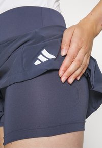 Pantalones cortos deportivos azul marino con una textura suave y una falda superpuesta. Presentan el logo blanco de Adidas y una cintura elástica para un ajuste seguro.