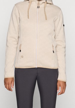 Sweat à capuche zippé beige texturé avec cordons et poches zippées, porté avec un pantalon foncé ajusté.