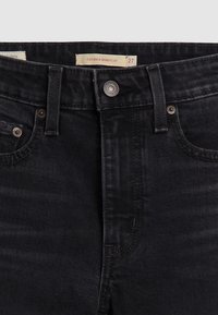Jean en denim noir, gros plan avant avec bouton, passants de ceinture, poches avant, et étiquette lisant "725 Kick Bootcut 27" visible à l'intérieur de la ceinture.