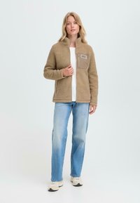 Femme portant une veste en polaire à fermeture éclair beige avec une poche poitrine, un t-shirt blanc, un jean bleu clair et des baskets beiges se tenant devant un fond uni.