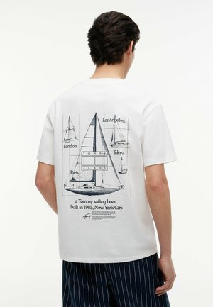 Mann trägt weißes T-Shirt mit schwarzer Segelboot-Grafik und Stadtnamen, darunter London, Paris, Los Angeles und Tokio, auf dem Rücken.