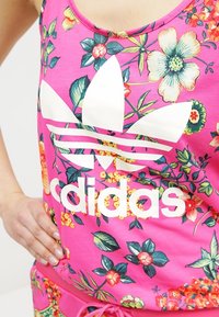 Ljust rosa blommönstrad linne med stort vitt Adidas-logotyp över bröstet, buren av en person med handen på höften.