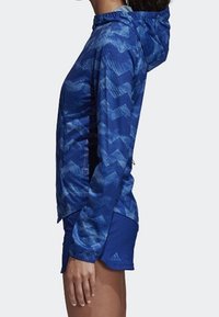 Veste légère bleue avec un motif en chevrons, dotée d'une capuche, d'une fermeture éclair à l'avant et d'un tissu texturé. Portée avec un short bleu.