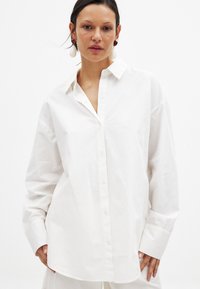 White button-up shirt made of smooth cotton. Features a classic collar, long sleeves, and a loose fit with subtle texture.
