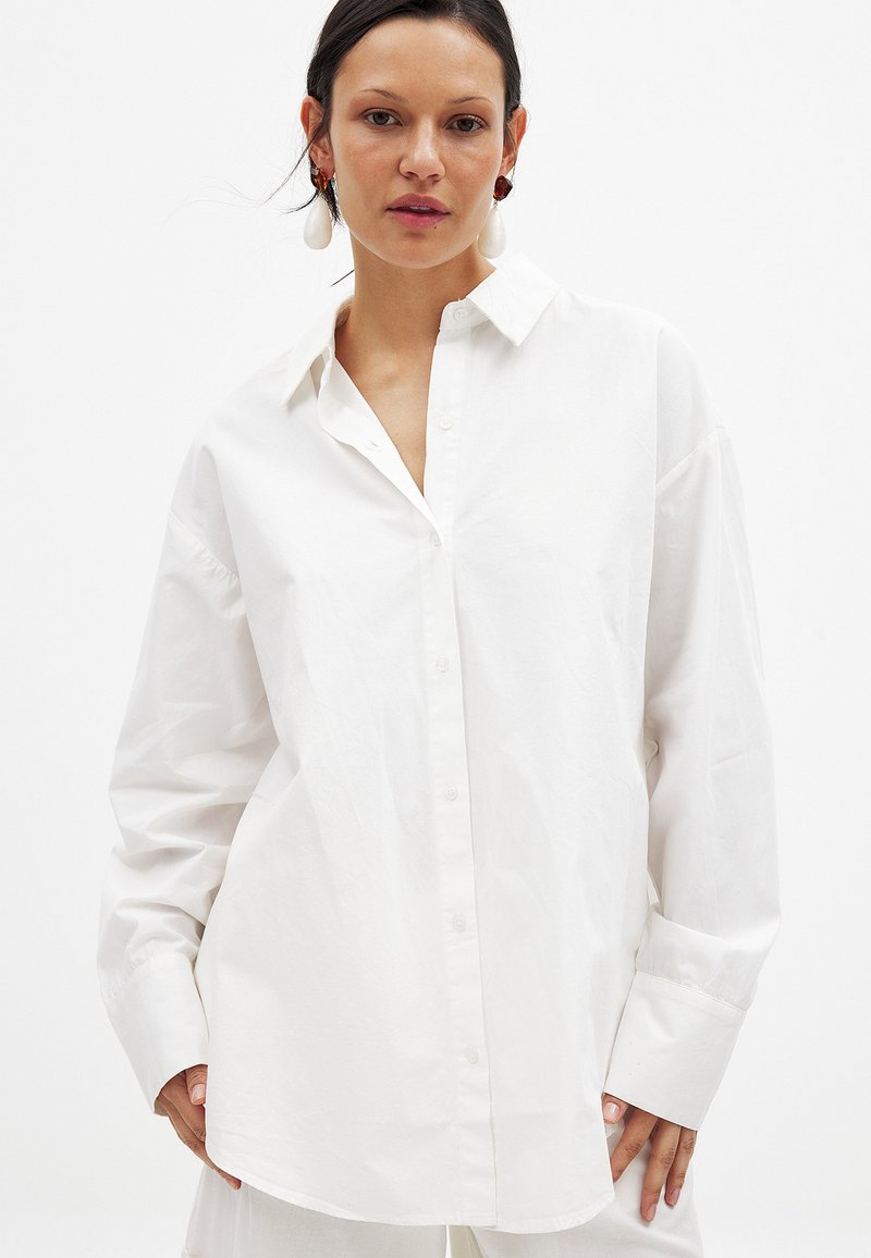 White button-up shirt made of smooth cotton. Features a classic collar, long sleeves, and a loose fit with subtle texture.