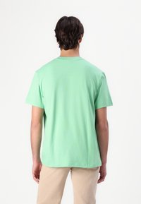 Personne aux cheveux courts et foncés portant un t-shirt vert clair à manches courtes et un pantalon beige, debout de dos sur un fond blanc.